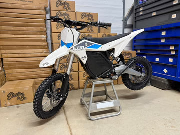 White dirt bike on a stand in a garage with boxes and a blue tool chest in the background.